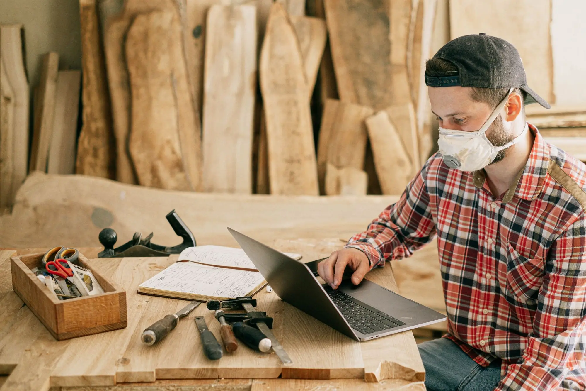 Joiner in a dust mask checking a new contract order on his laptop at a timber workbench in his workshop